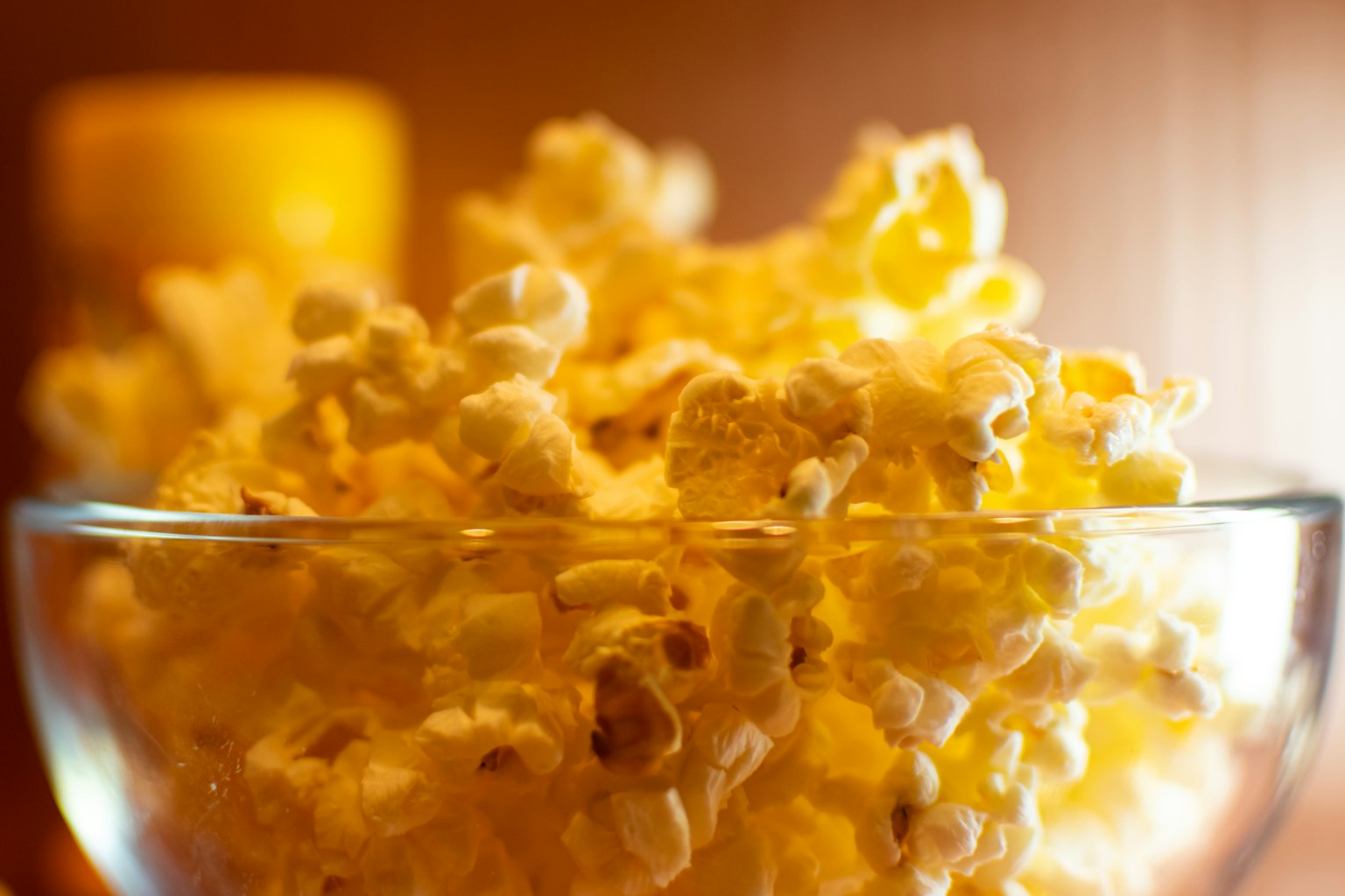A glass bowl heaped with warm golden popcorn, lit by soft evening light beside a candle