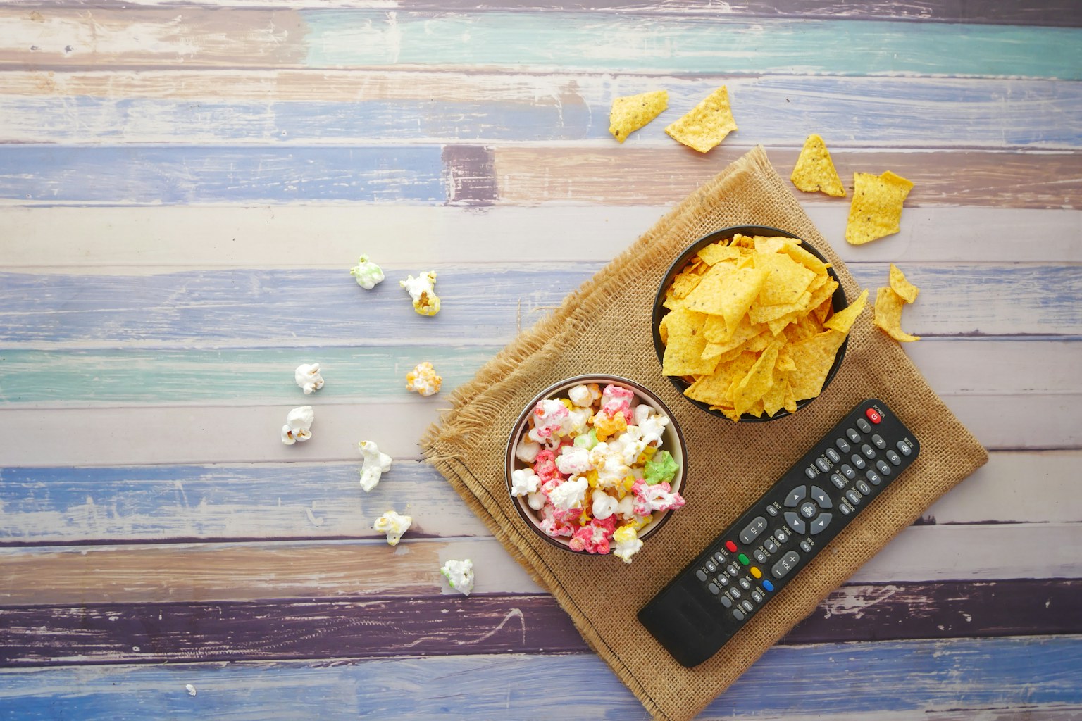 A bowl of colourful popcorn and a bowl of corn chips on a hessian cloth beside a TV remote